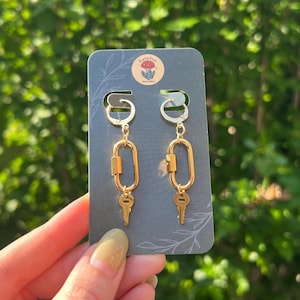 Op de afbeelding: Goudkleurige oorbellen met een sleutel- en slotontwerp. De oorbellen hangen aan een donkergrijze kaart met de tekst "Earth Star" en een bloemenlogo. De achtergrond is een wazig groen.