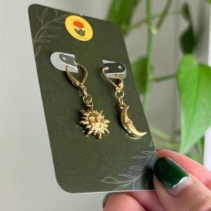 Puede incluir: Pendientes dorados con un sol y una luna creciente. Los pendientes se exhiben en una tarjeta verde oscuro con una peque&ntilde;a pegatina y un dise&ntilde;o de hojas. Los pendientes son sostenidos por una persona con esmalte de u&ntilde;as verde.