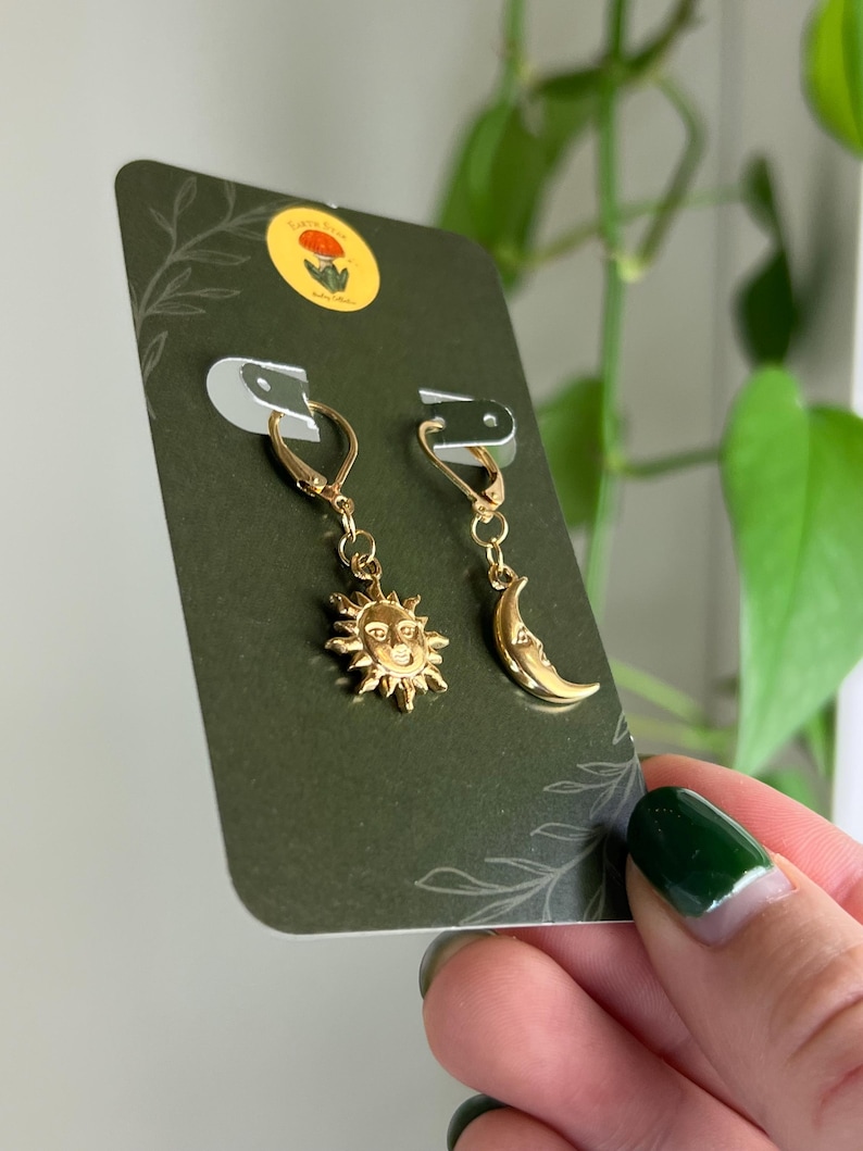 Puede incluir: Pendientes dorados con un sol y una luna creciente. Los pendientes se exhiben en una tarjeta verde oscuro con una peque&ntilde;a pegatina y un dise&ntilde;o de hojas. Los pendientes son sostenidos por una persona con esmalte de u&ntilde;as verde.