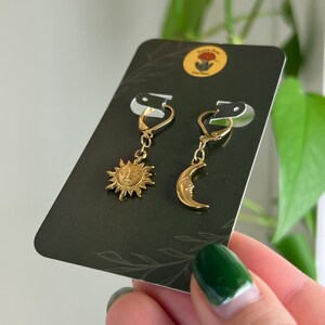 Puede incluir: Pendientes celestiales dorados, uno con un sol y otro con una luna creciente. El sol tiene una cara detallada, y la luna tiene una cara sutil. Los pendientes se muestran en una tarjeta verde oscuro con un peque&ntilde;o logotipo.