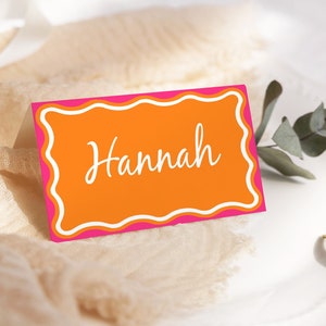 May include: Orange and pink place card with a white scalloped border and the name "Hannah" written in white cursive.