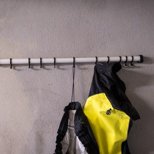 Könnte beinhalten: Ein weißer Metall-Kleiderständer mit schwarzen Haken, der an einer grauen Wand montiert ist. Zwei Jacken hängen an den Haken, eine schwarze und eine gelbe.