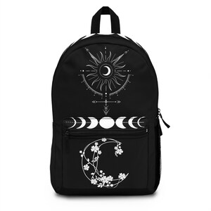 May include: Black backpack featuring a celestial design with a sun, moon phases, and floral crescent moon. The backpack has a zippered main compartment, a front zippered pocket, and a top handle. The design is in white against a black background.