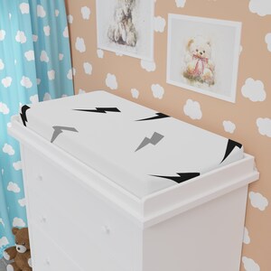 May include: A white changing pad cover with a pattern of black and gray lightning bolts. The cover is placed on a white changing table. The background includes a light blue cloud-patterned curtain and framed teddy bear art.
