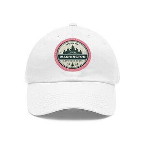 May include: White baseball cap with a circular patch. The patch reads "BORN IN PNW WASHINGTON LOVE YOUR ROOTS 1967" with a forest graphic. The patch has a pink border and a cream background.