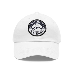 May include: White baseball cap with a circular patch featuring the text "Washington Beautiful Day" and "PNW Roots" around a mountain graphic. The cap has a curved peak and an adjustable design.