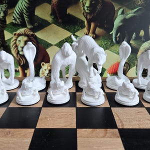 Può includere: Un originale set di scacchi con pezzi bianchi a forma di animali, tra cui leoni, cobra e gorilla, esposti su una classica scacchiera a scacchi neri e marroni. Lo sfondo mostra ulteriori figurine di animali.
