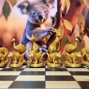 May include: A gold-colored chess set featuring animal-shaped pieces, including kangaroos, emus, and wombats, arranged on a checkered board. The background shows a koala and foliage. This unique set is perfect for chess enthusiasts.