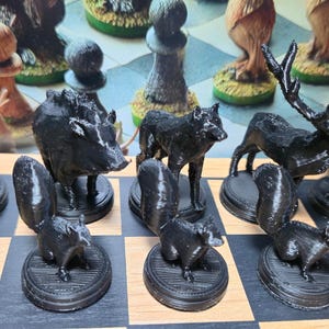 K&ouml;nnte beinhalten: Ein schwarzes Schachspiel mit Tierfiguren. Das Set enth&auml;lt Figuren in Form eines Eichh&ouml;rnchens, Wolfs, Wildschweins und Hirsches, die auf einem Schachbrett angeordnet sind. Die Figuren sind detailliert und heben sich von den hellen und dunklen Feldern ab.