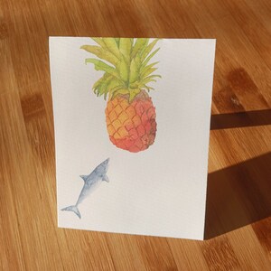 Könnte beinhalten: Ein Aquarellbild von einer Ananas mit einem blauen Hai, der darunter schwimmt. Die Ananas ist gelb und orange mit grünen Blättern. Der Hai ist hellblau.