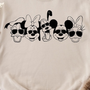 Könnte beinhalten: Ein beiger Pullover mit einer schwarzen Grafik von Mickey Mouse, Minnie Mouse, Donald Duck, Goofy und Pluto, die Sonnenbrillen tragen.