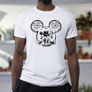 May include: White t-shirt featuring a black and white illustration of Mickey and Minnie Mouse. The design includes a large outline of Mickey's head with fireworks and stars. The cartoon couple is depicted sitting together.