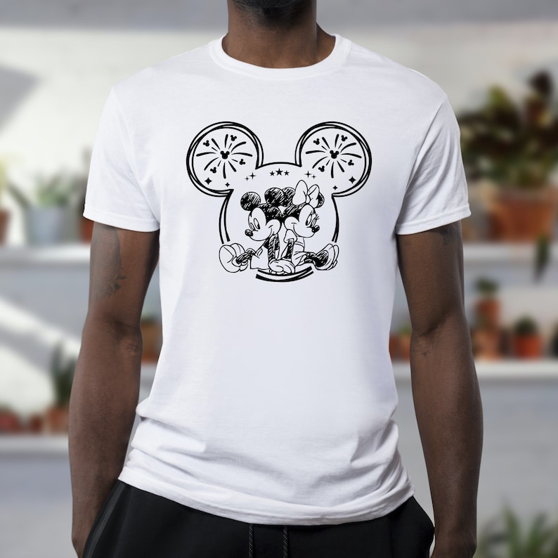 May include: White t-shirt featuring a black and white illustration of Mickey and Minnie Mouse. The design includes a large outline of Mickey's head with fireworks and stars. The cartoon couple is depicted sitting together.