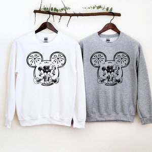 May include: Two sweatshirts, one white and one gray, each featuring a black and white graphic of Mickey and Minnie Mouse. The design includes Mickey Mouse ears with fireworks and the couple embracing. The sweatshirts are hanging on wooden hangers.