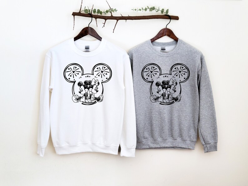 May include: Two sweatshirts, one white and one gray, each featuring a black and white graphic of Mickey and Minnie Mouse. The design includes Mickey Mouse ears with fireworks and the couple embracing. The sweatshirts are hanging on wooden hangers.