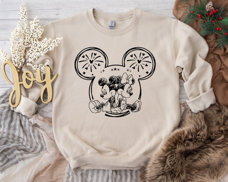 May include: Beige sweatshirt featuring a black and white illustration of Mickey and Minnie Mouse. The design includes fireworks and stars within the iconic mouse ears. The sweatshirt is a crew neck style.
