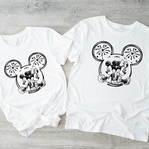 May include: Two white t-shirts featuring a black and white graphic of Mickey and Minnie Mouse. The design includes a Mickey Mouse head outline with fireworks and the couple sitting back-to-back. The shirts are short-sleeved.