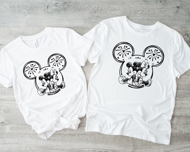May include: Two white t-shirts featuring a black and white graphic of Mickey and Minnie Mouse. The design includes a Mickey Mouse head outline with fireworks and the couple sitting back-to-back. The shirts are short-sleeved.