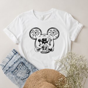 May include: White t-shirt featuring a black and white graphic of Mickey and Minnie Mouse embracing. The design is set within a Mickey Mouse ear outline, with fireworks and stars. The shirt is paired with denim shorts and a straw hat.