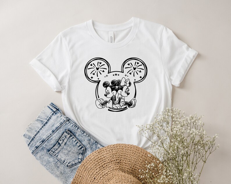 May include: White t-shirt featuring a black and white graphic of Mickey and Minnie Mouse embracing. The design is set within a Mickey Mouse ear outline, with fireworks and stars. The shirt is paired with denim shorts and a straw hat.