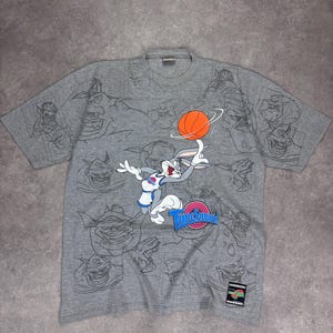 May include: Grey short-sleeved t-shirt featuring Bugs Bunny playing basketball with the text "Tune Squad". The shirt has a collage of cartoon character sketches. A small tag is visible on the bottom right.