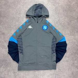 Könnte beinhalten: Grauer Kapuzenpullover mit Reißverschluss und blauen Akzenten sowie einem Teamlogo. Die Jacke hat blaue Streifen auf den Ärmeln und der Kapuze sowie ein weißes Logo auf der Brust. Die Ärmel sind marineblau.