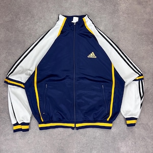 Puede incluir: Chaqueta deportiva Adidas azul marino con detalles blancos y amarillos. La chaqueta tiene cremallera completa, dos bolsillos delanteros y el logo de Adidas. Las mangas tienen tres rayas negras.