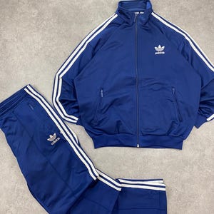 Könnte beinhalten: Ein marineblaues Trainingsanzug-Set mit einer Jacke mit Reißverschluss und passender Hose. Jacke und Hose haben weiße Streifen an den Seiten und das Adidas-Logo. Der Trainingsanzug ist aus glänzendem Material.