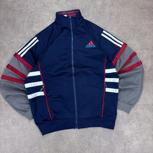 Könnte beinhalten: Eine marineblaue Adidas-Trainingsjacke mit durchgehendem Reißverschluss. Die Jacke hat weiße Streifen auf den Ärmeln und rote Akzente. Das Adidas-Logo ist rot auf der Brust. Die Ärmel sind grau.