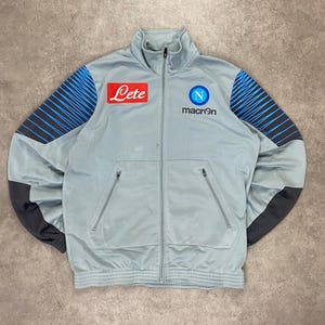 Könnte beinhalten: Hellgraue Trainingsjacke mit blauen und schwarzen Akzenten. Sie hat einen durchgehenden Reißverschluss, zwei Vordertaschen und die Logos "Lete", "macron" und ein blaues "N"-Logo. Die Ärmel haben blau-schwarze Streifenmuster.
