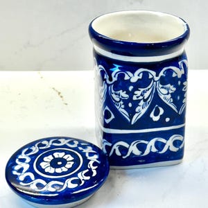 May include: A blue and white ceramic jar with a matching lid. The jar and lid feature a detailed floral and scroll design. The jar is cylindrical with a slightly flared rim. The lid is round and flat, with a central floral motif.