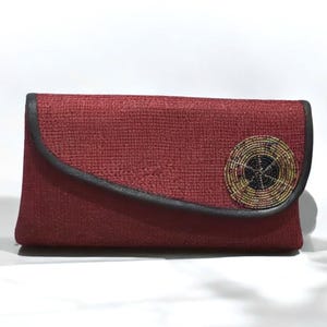 May include: A burgundy clutch with a textured fabric. The flap is outlined with black trim and features a circular beaded embellishment with gold, black, and yellow beads. The clutch is rectangular in shape.
