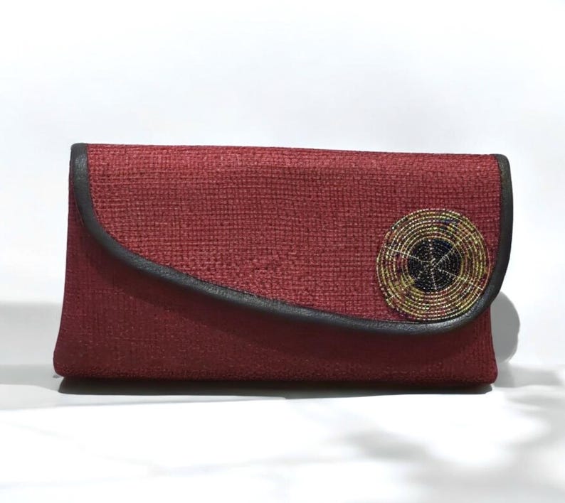 May include: A burgundy clutch with a textured fabric. The flap is outlined with black trim and features a circular beaded embellishment with gold, black, and yellow beads. The clutch is rectangular in shape.