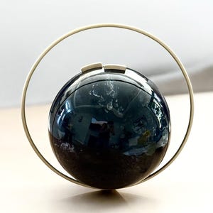 May include: A dark, spherical object with a reflective surface, held within a circular, gold-colored frame. Two small, rectangular accents sit atop the sphere. The object's surface reflects the surrounding environment.