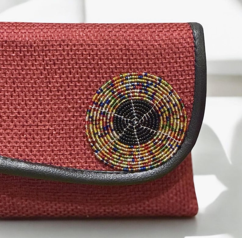 May include: A red woven clutch with a circular beaded embellishment. The beaded design features concentric circles of black, yellow, blue, green, and white beads. The clutch has a black leather trim.