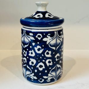 May include: A ceramic jar with a lid, featuring a dark blue base and white floral patterns. The jar has a cylindrical shape with a matching lid. The design includes white leaf and dot patterns on a dark blue background. The lid has a white knob.