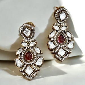 May include: A pair of gold tone dangle earrings with white and red stones. The earrings have a floral design and are accented with small clear stones.