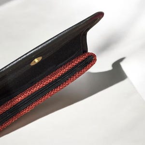 May include: Open black leather clutch with a red woven zipper pocket. The interior is black, and a gold-colored snap closure is visible. The clutch is partially open, revealing the interior.