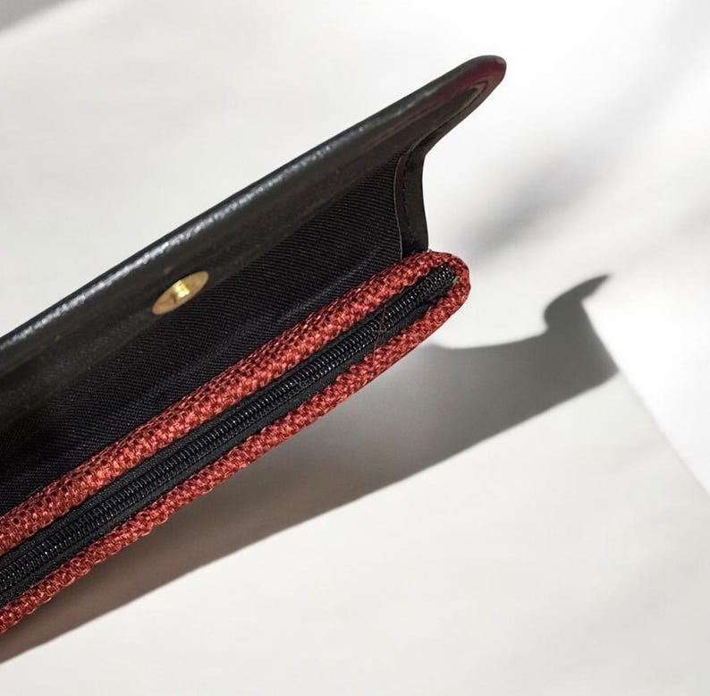 May include: Open black leather clutch with a red woven zipper pocket. The interior is black, and a gold-colored snap closure is visible. The clutch is partially open, revealing the interior.