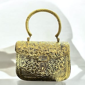 May include: A gold-colored, ornate handbag with a curved handle. The bag features intricate floral and scrollwork patterns across its surface, with a clasp closure. The handle is a semi-circular shape, and the bag has a rectangular body.