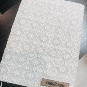 May include: A white, rectangular fabric item with an embroidered floral pattern. The design features a repeating diamond and flower motif. A small, rectangular brown tag with the text "FABRICLORE" is attached to the fabric.