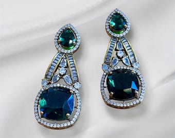 May include: A pair of elegant drop earrings featuring large, square-cut, dark green gemstones. The earrings have a gold-tone setting with a halo of small, clear stones and a teardrop-shaped green stone at the top.