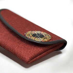 May include: A rectangular, rust-colored clutch with a woven texture. The clutch features a black leather trim and a circular, beaded embellishment in a variety of colors. The clasp is hidden under the flap.