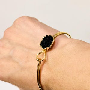 May include: Gold-tone bangle bracelet featuring a black, faceted, hexagonal stone. The stone is set in a gold-colored bezel. The bracelet has a simple, elegant design, suitable for everyday wear or special occasions.