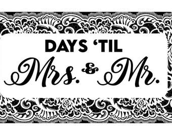 May include: A rectangular sign with a white background and black decorative border. The text "DAYS 'TIL Mrs. & Mr." is displayed in black, with the words "Mrs. & Mr." in an elegant script font. The border features a detailed floral design.