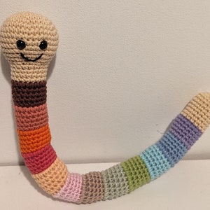 Op de afbeelding: Een gehaakt wormpje met een glimlachend gezicht en een veelkleurig lichaam. De worm is gemaakt van verschillende tinten bruin, oranje, roze en blauw garen.