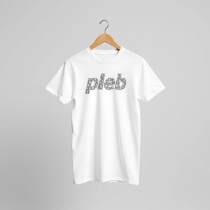 Puede incluir: Camiseta blanca con la palabra "pleb" impresa en gris en el pecho. Las letras están rellenas con un patrón de pequeños símbolos de Bitcoin. La camiseta está colgada en una percha de madera.