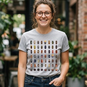 Puede incluir: Una camiseta de manga corta en color gris jaspeado con un patrón de cuadrícula que muestra varias ilustraciones coloridas de cochinillas. El gráfico presenta múltiples filas de pequeños crustáceos ovalados en tonos marrones, grises, azules y naranjas.