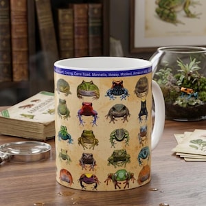 May include: White ceramic mug featuring a collage of colorful frog illustrations. The mug has a cream-colored background with various frog species. The top of the mug has a blue band with text. A book and magnifying glass are in the background.
