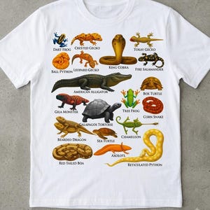 Könnte beinhalten: Ein weißes kurzärmeliges T-Shirt mit einem farbenfrohen Grafikdruck verschiedener Reptilien und Amphibien, darunter eine Königskobra, ein Mississippi-Alligator und eine Galápagos-Riesenschildkröte, jeweils mit ihrem Namen in schwarzem Text.